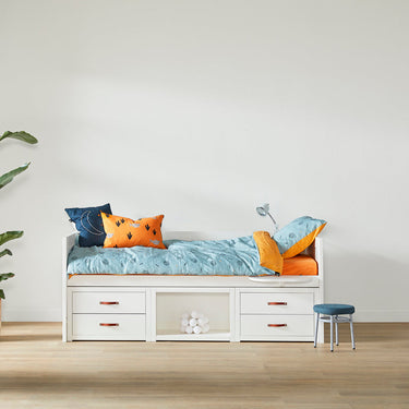 White bed with colorful bedding in a minimalistic room with a plant and stool.